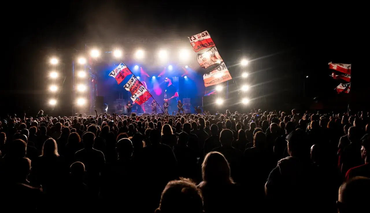Tłum ludzi na koncercie podczas Cieszanów Festiwal. Na scenie zespół gra, a nad głowami publiczności powiewają flagi.