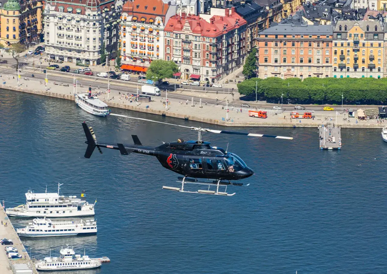 En svart helikopter flyger &ouml;ver vattnet i Stockholm. I bakgrunden syns byggnader och b&aring;tar.