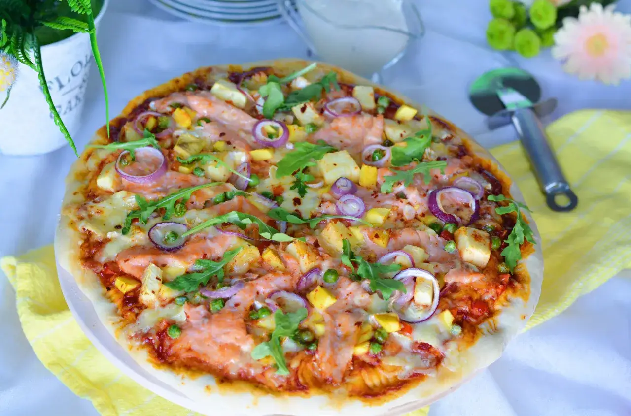 Pyszna pizza z łososiem, ananasem, cebulą i rukolą, posypana zielonym groszkiem i płatkami chili.