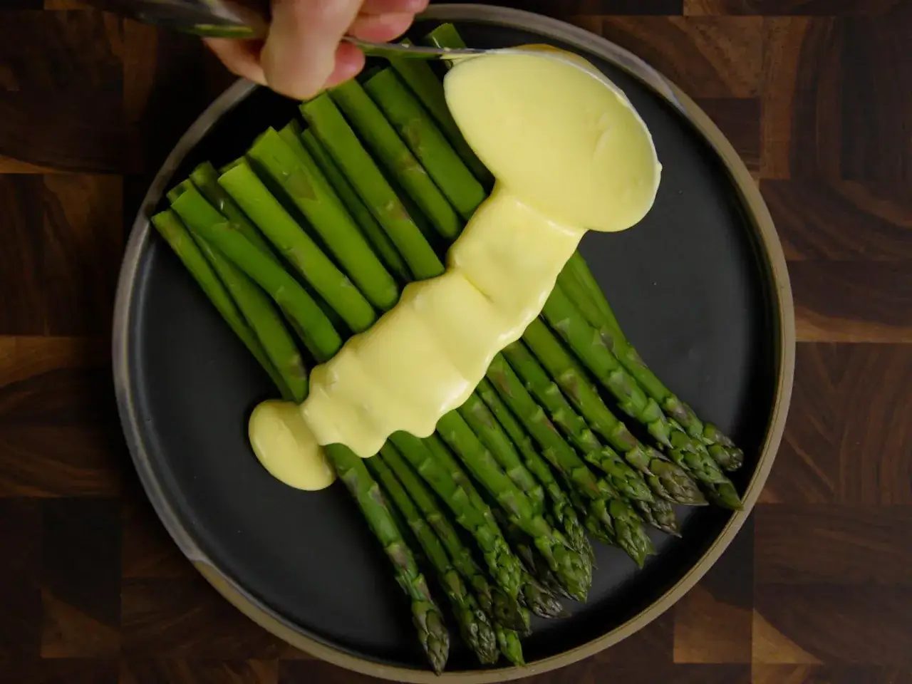 Une main verse une sauce hollandaise onctueuse sur une botte d'asperges vertes sur une assiette sombre.