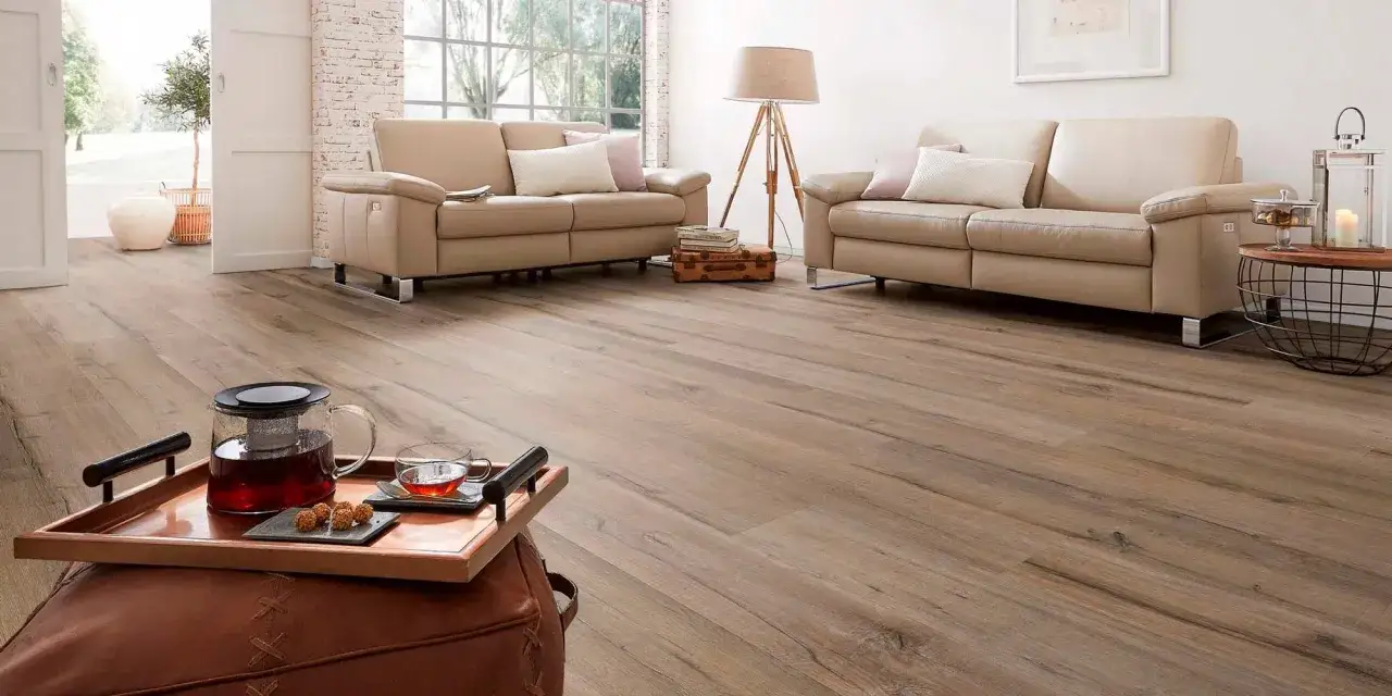 Un salón acogedor con sofás beige y un suelo laminado o parquet de madera clara. Una bandeja con té y dulces sobre un puf de cuero.