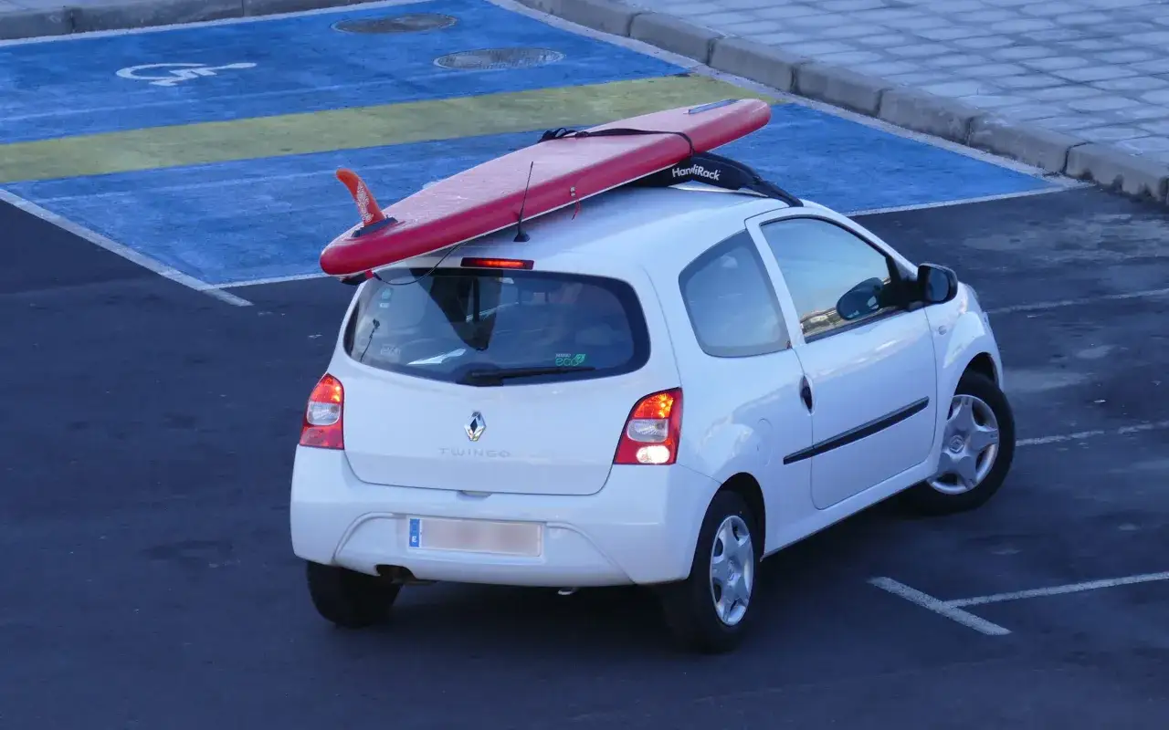 Wei&szlig;er Renault Twingo mit rotem Surfbrett auf dem Dach, bereit, um Wellen zu transportieren.