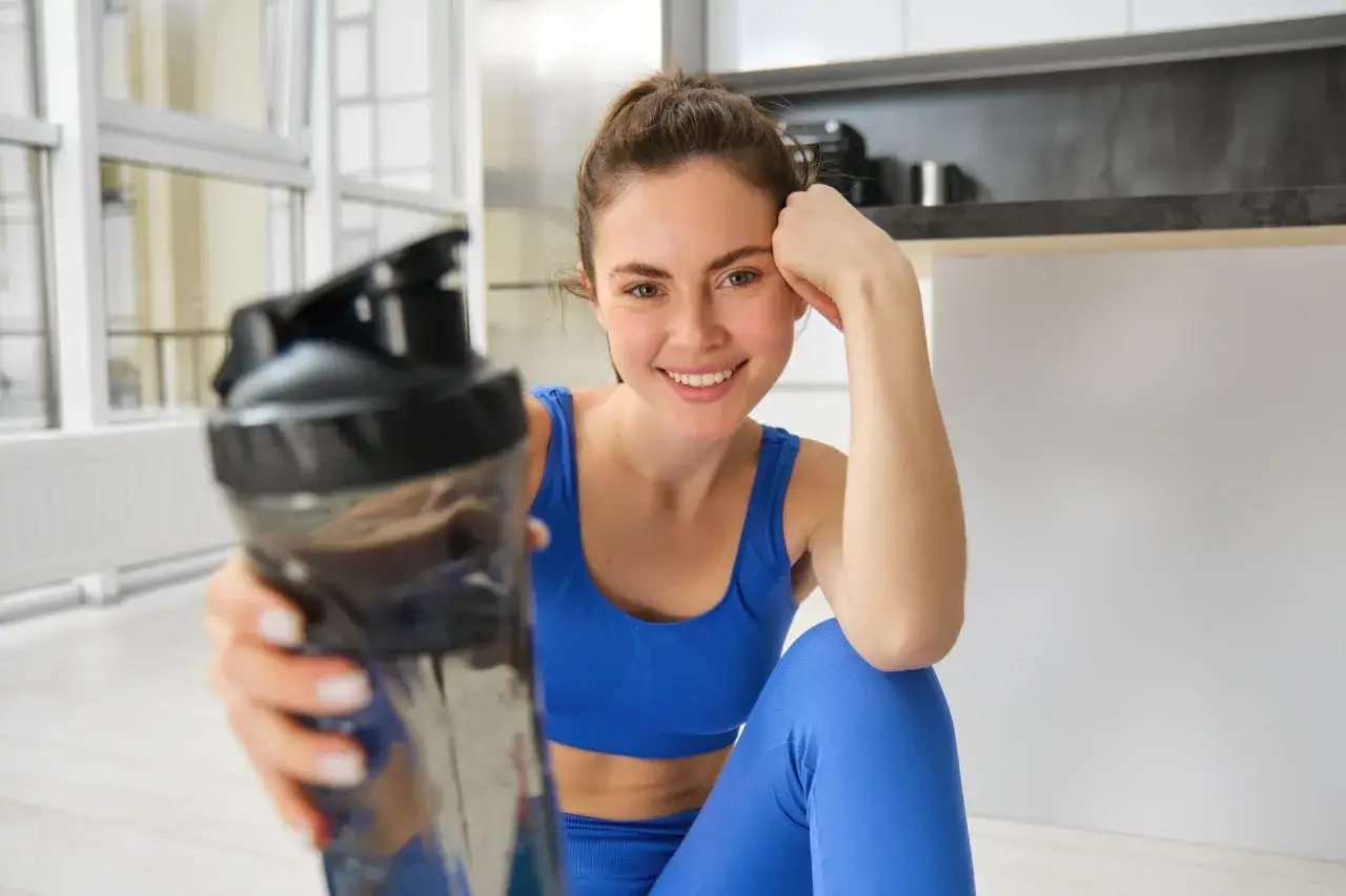 Uśmiechnięta kobieta w niebieskim stroju sportowym trzyma shaker z napojem. Czy to białko przed czy po treningu?