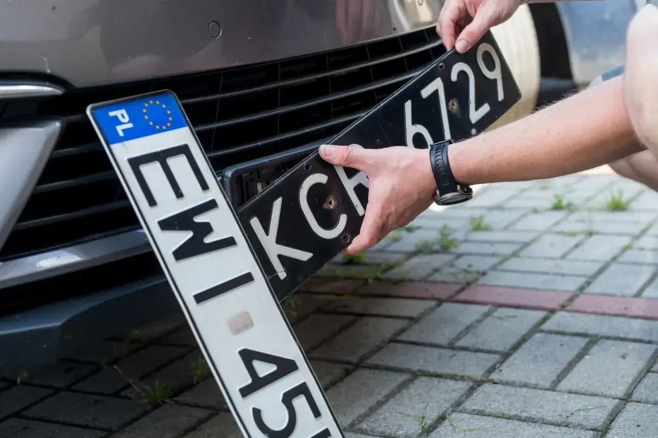 Wymiana tablic rejestracyjnych. Jedna tablica z PL i unijnymi gwiazdkami, druga czarna z białymi znakami.