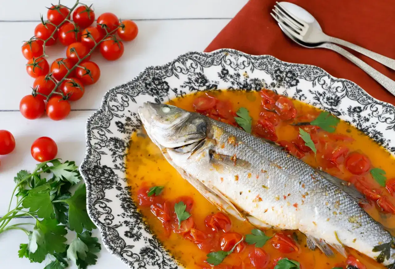 Spigola all'acqua pazza in un piatto decorato, con pomodorini e prezzemolo fresco.