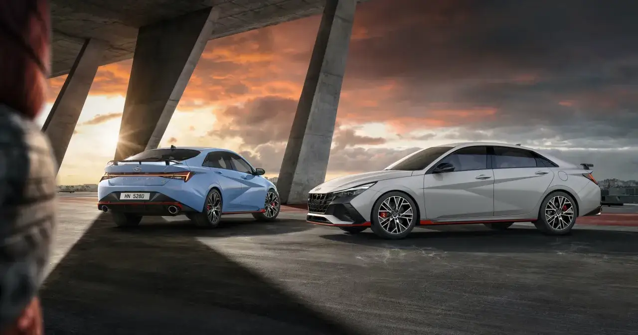 Dwa sportowe samochody Hyundai Elantra N, jeden niebieski, drugi jasnoszary, stoją pod betonowym mostem o zachodzie słońca.