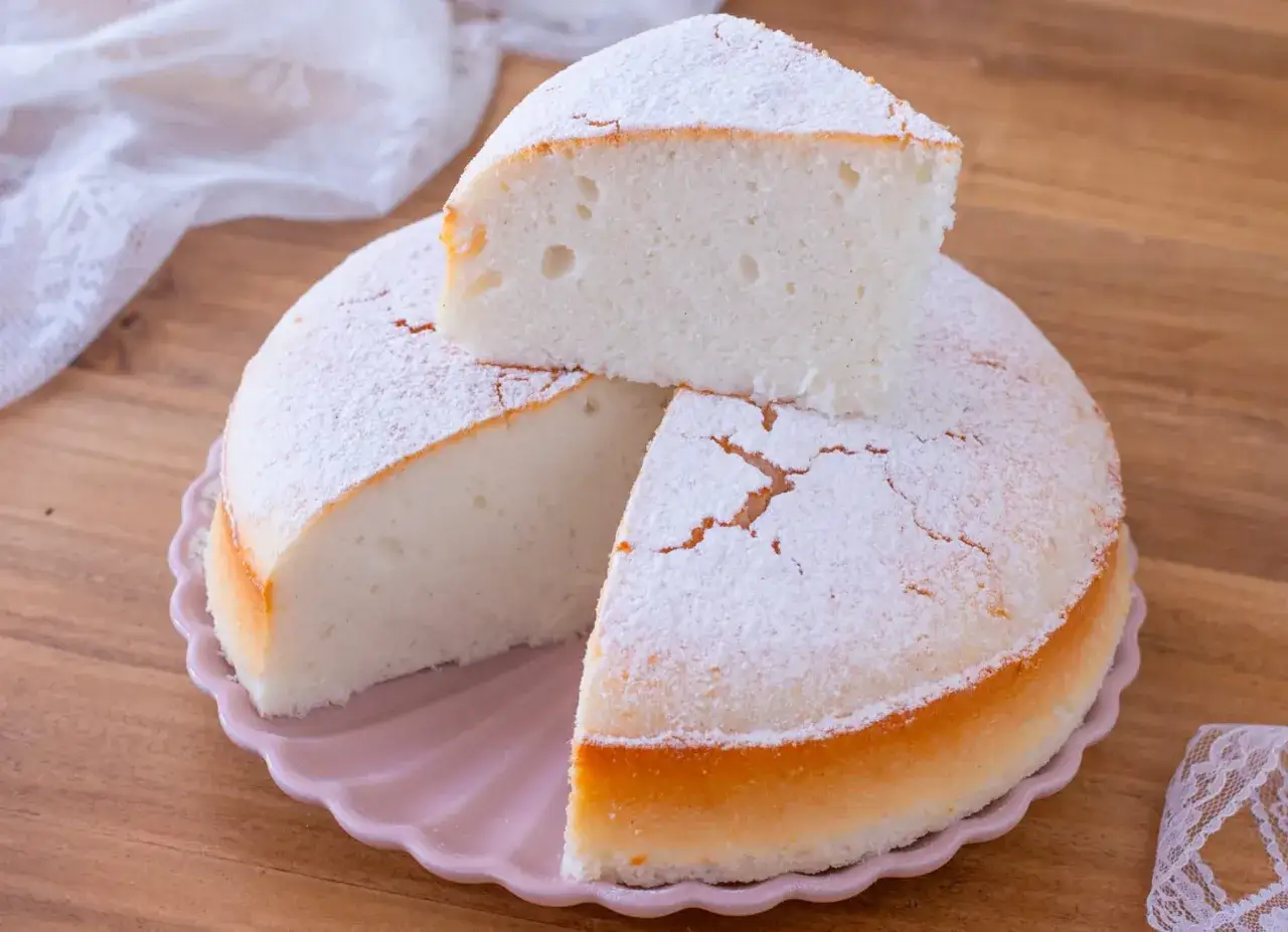 Una soffice torta nuvola, cosparsa di zucchero a velo, su un piatto rosa. Una fetta &egrave; appoggiata sopra, invitando a gustarla.