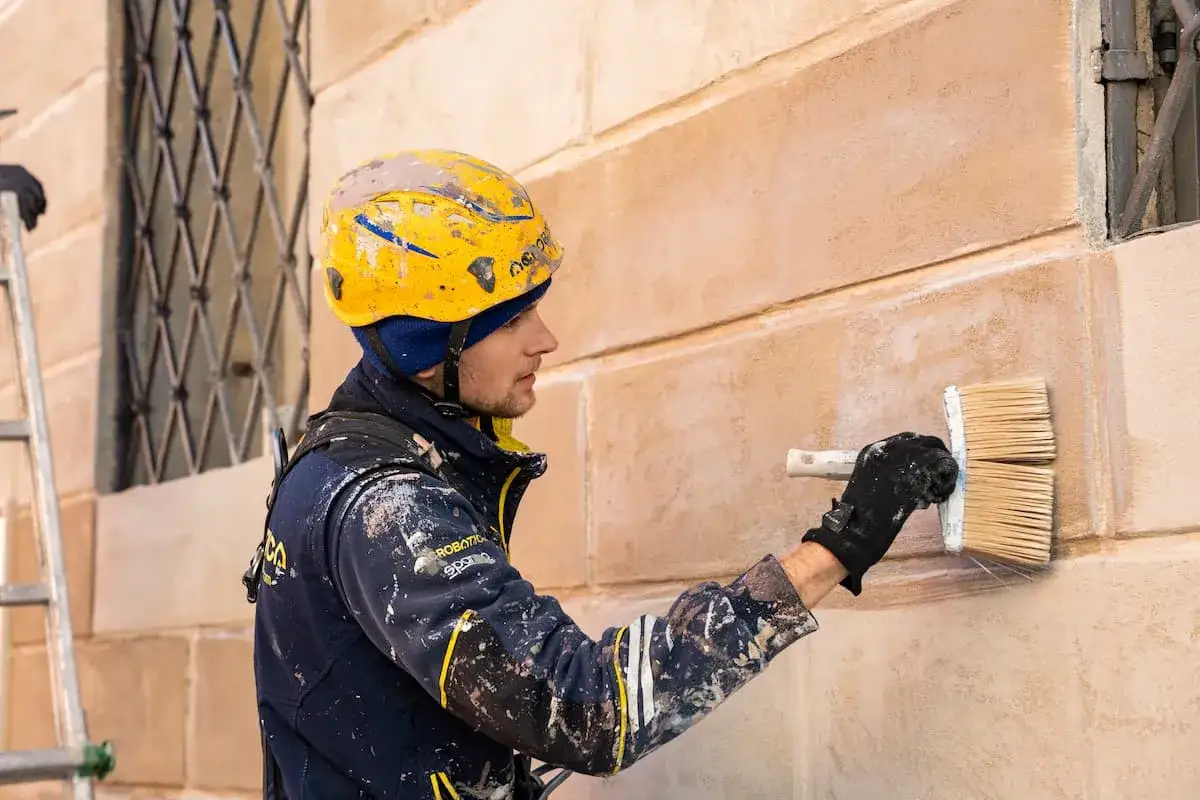 Operaio con casco giallo intento a intonacare muro vecchio con un pennello largo.