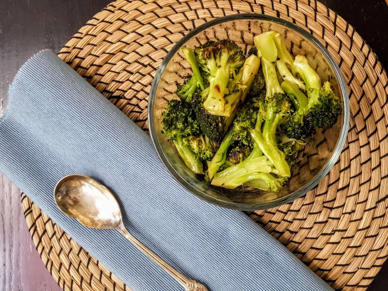 Brotes de br&oacute;coli asado en un taz&oacute;n de vidrio, listos para ser parte de tus recetas con br&oacute;coli. Servilleta azul y cuchara antigua al lado.
