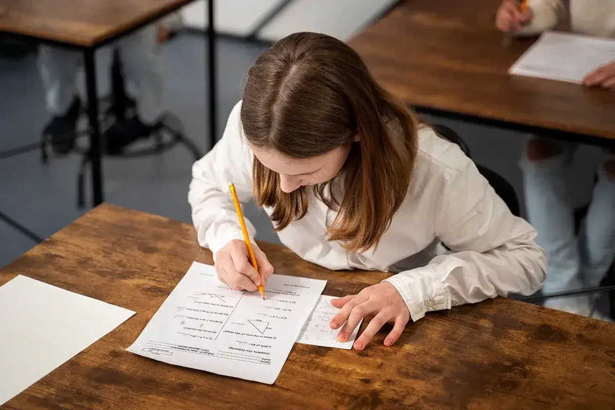 Dziewczynka pisze coś na kartce z zadaniami matematycznymi, siedząc przy drewnianym biurku.