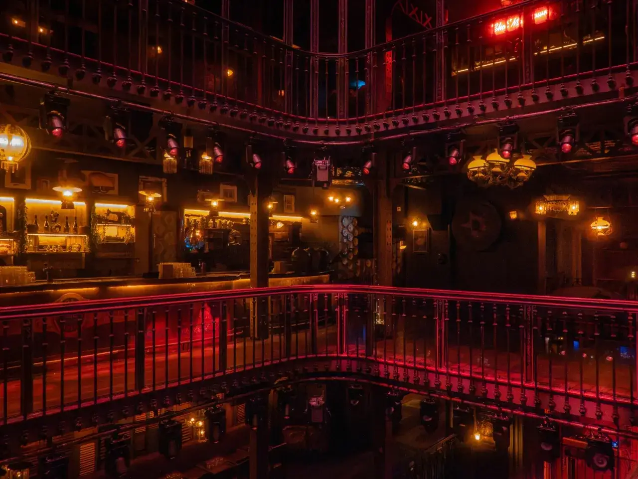 L'int&eacute;rieur d'un bar &agrave; l'ambiance festive, &eacute;clair&eacute; par des lumi&egrave;res rouges et dor&eacute;es. On aper&ccedil;oit un bar, des balustrades orn&eacute;es et des lustres, &eacute;voquant un **barrio latino** &agrave; Paris.
