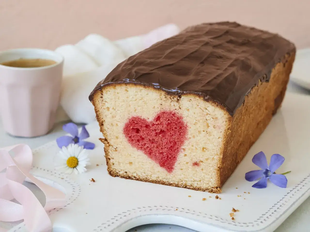 Schokoladen&uuml;berzogener Kastenkuchen mit einem roten Herz im Inneren, serviert mit Kaffee und Blumen.