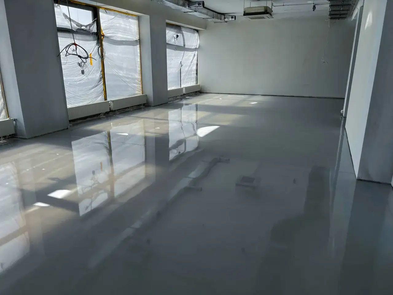Nowe, lśniące posadzki z żywicy w pustym pomieszczeniu. Okna zasłonięte folią, widoczne instalacje.