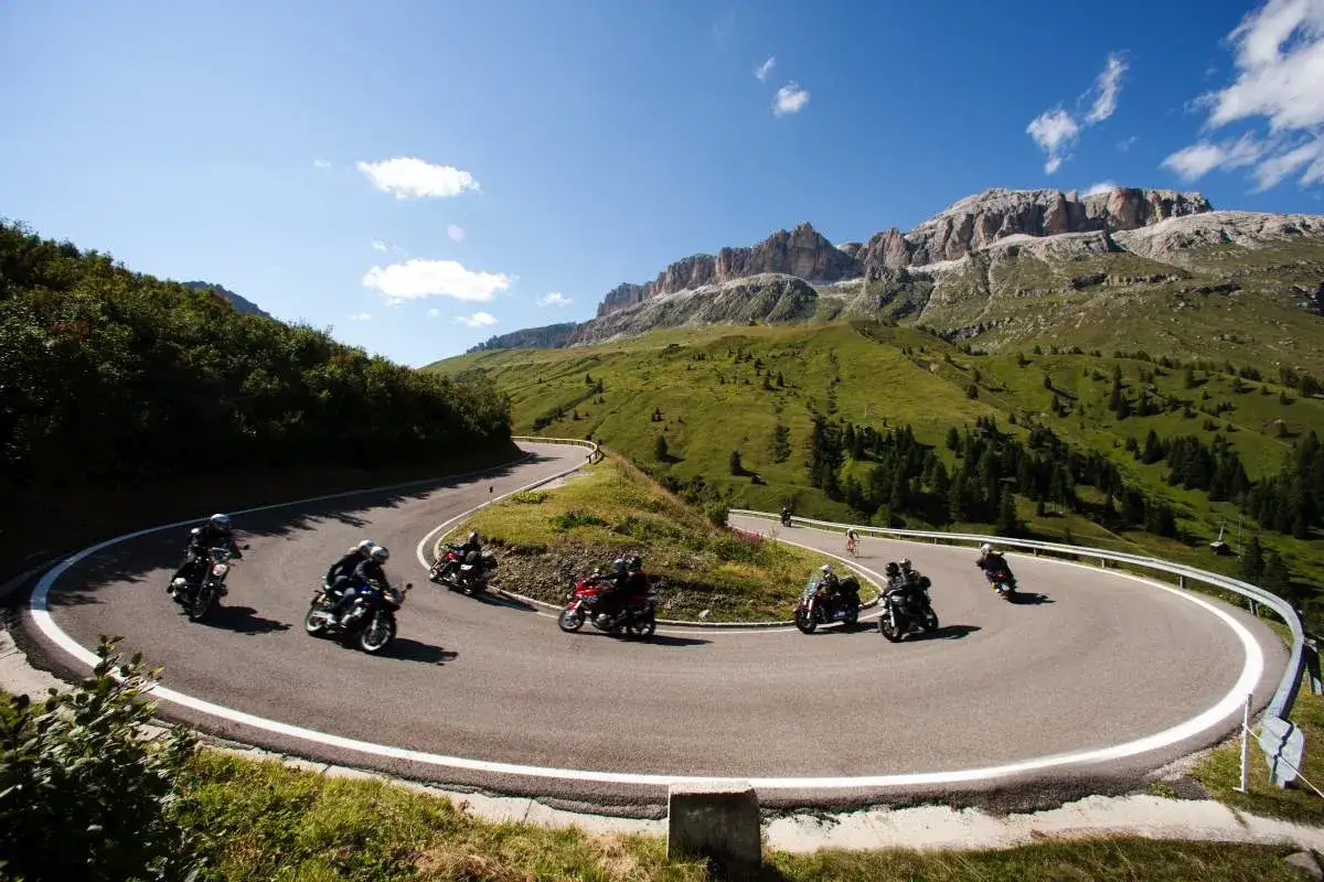 Motociclisti in gruppo affrontano un tornante su una strada panoramica, con montagne verdi e rocciose sullo sfondo. Un'avventura sul passo selva.