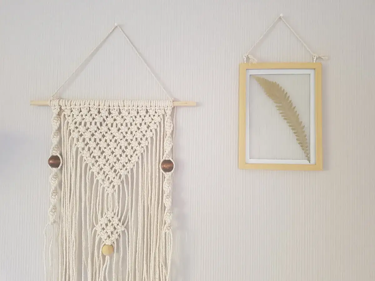 Décoration murale macramé tuto : un artisanat en cordelette écru avec perles en bois, suspendu à côté d'un cadre jaune contenant une fougère séchée.