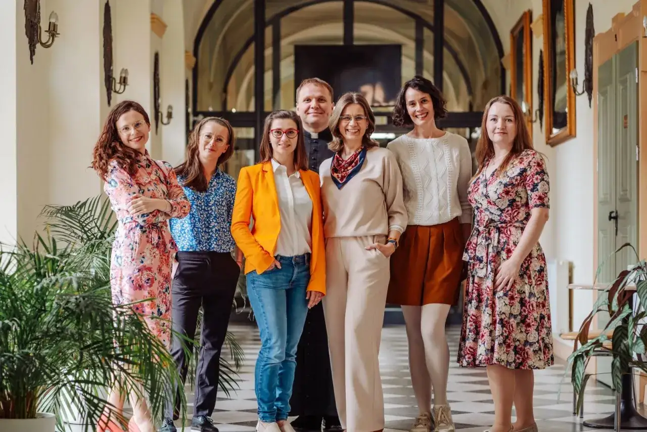 Grupa osób, w tym ksiądz, omawia adopcja dziecka warunki. Uśmiechnięci, stoją w eleganckim korytarzu.