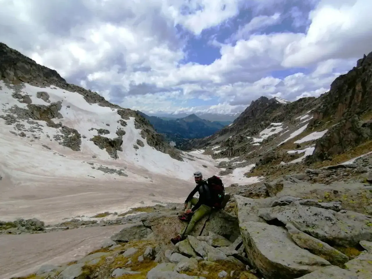 Un excursionista descansa en el **valle de Aneu**, rodeado de picos rocosos y parches de nieve bajo un cielo nublado.