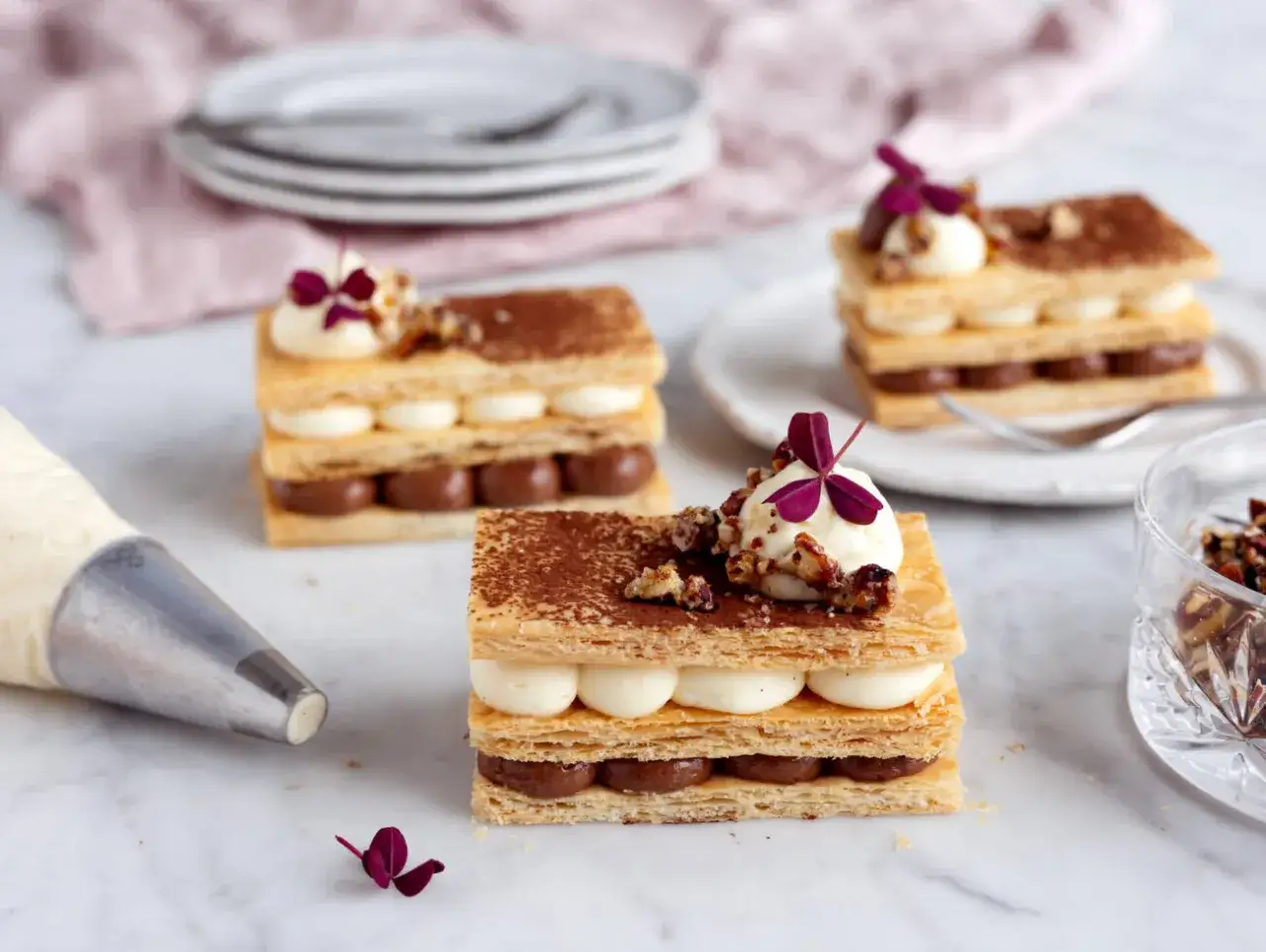 Tre eleganta millefeuille-desserter med lager av krispigt bakverk, chokladkräm och vaniljkräm, toppade med nötter och en lila blomma.