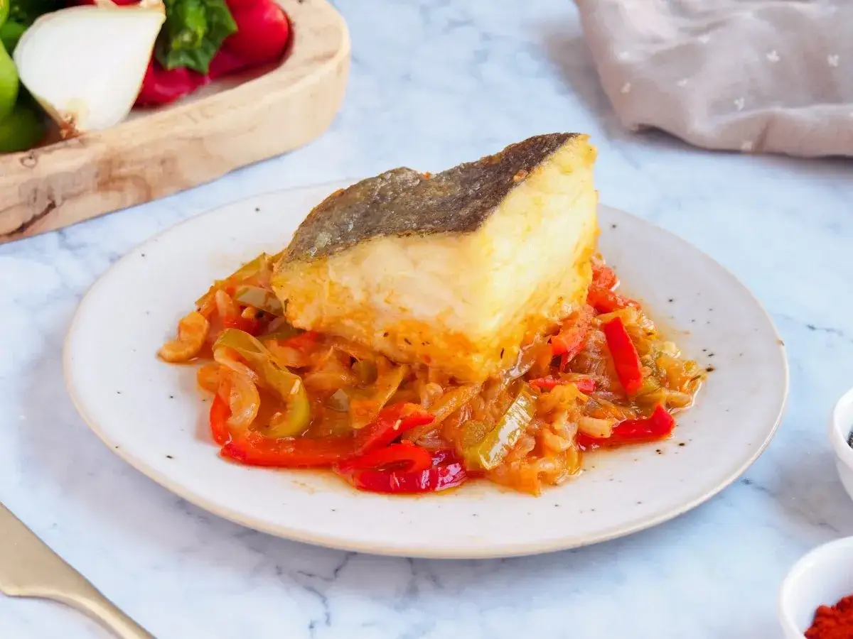 Un trozo de bacalao encebollado reposa sobre una cama de pimientos y cebolla.