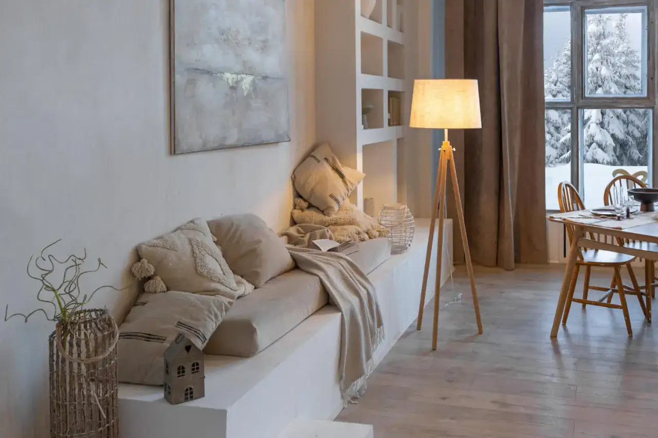 Un rincón acogedor con cojines y manta, lámpara de pie de madera y ventana nevada. Puro minimalismo cálido.