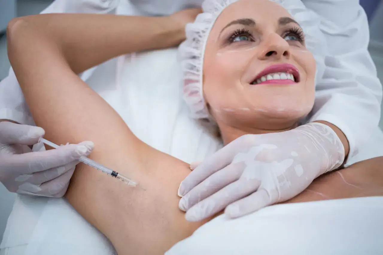 Kobieta z uśmiechem na twarzy poddaje się zabiegowi botox pod pachy. Lekarz w rękawiczkach wykonuje zastrzyk.
