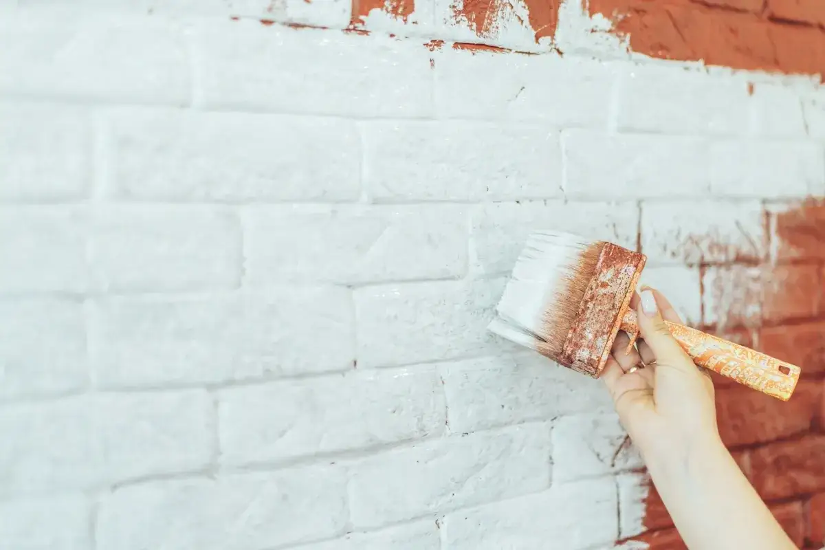 Application d'enduit sur brique int&eacute;rieur. Une main tient un gros pinceau pour peindre un mur de briques en blanc.