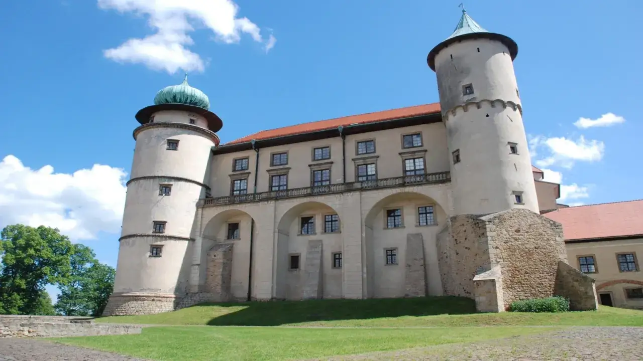 Zamek Wiśnicz z dwiema wieżami i zielonymi kopułami, otoczony zielenią, pod błękitnym niebem z chmurami.