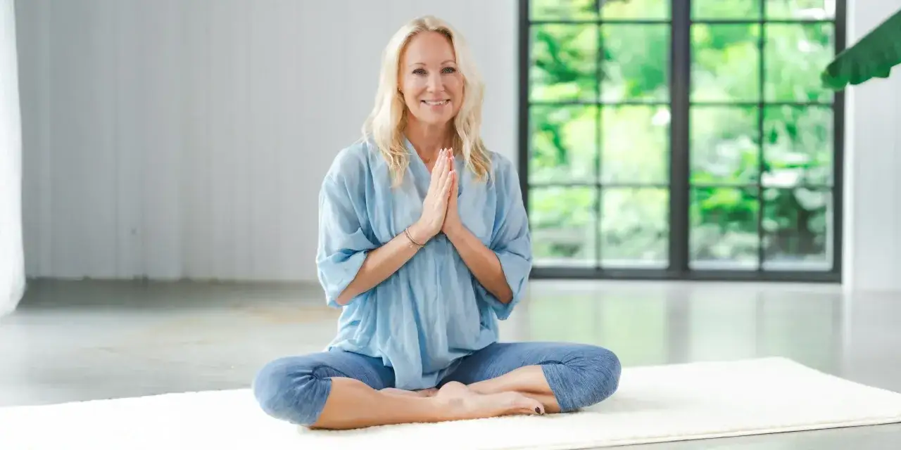 Kvinna med blont h&aring;r sitter i meditationsst&auml;llning med h&auml;nderna i b&ouml;n. Hon ler mot kameran, kl&auml;dd i en ljusbl&aring; skjorta och bl&aring; byxor. Bakom henne syns ett stort f&ouml;nster med gr&ouml;nska. Perfekt f&ouml;r en guidad meditation text.