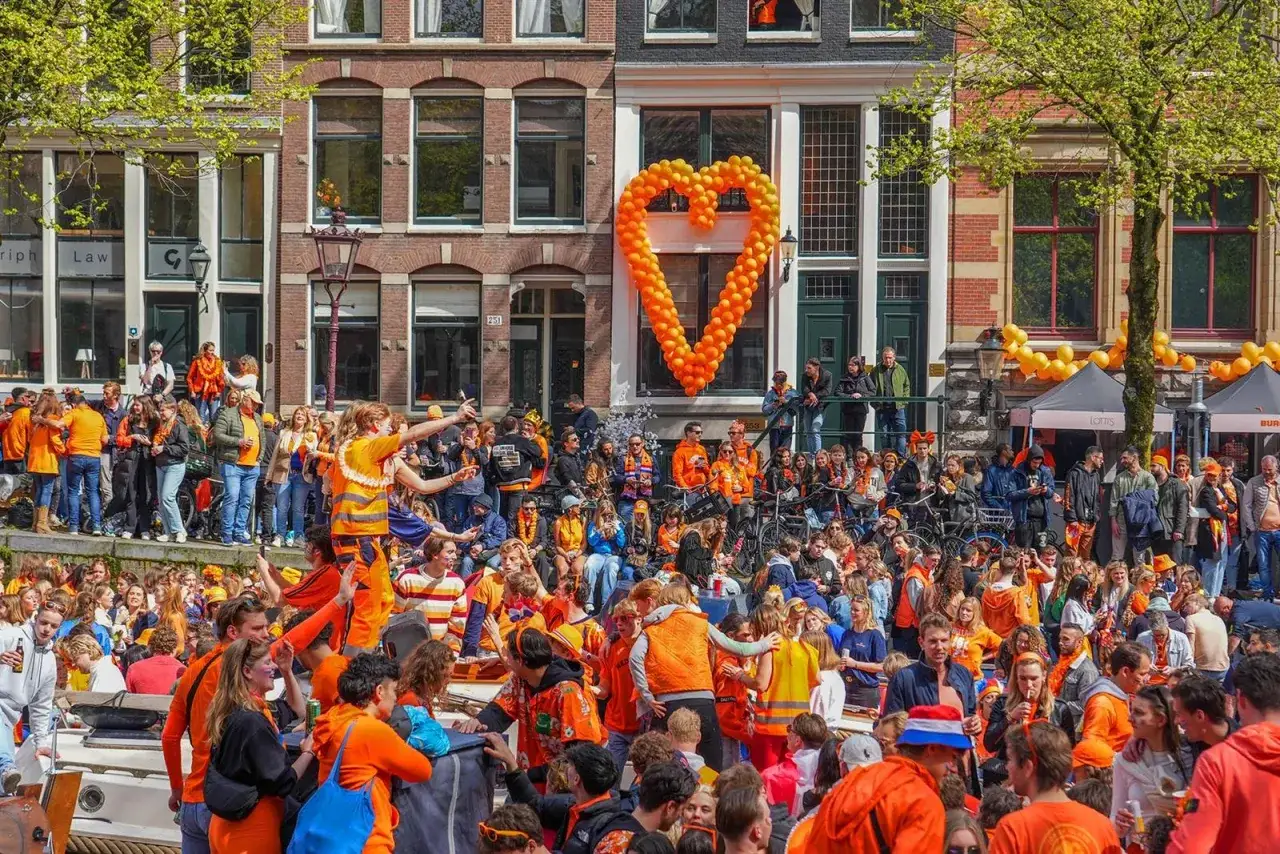 Grote oranje hartenballonnen sieren de grachten van Amsterdam tijdens een feestelijke dag. Mensen in oranje kleding vieren feest op boten en langs de kades.