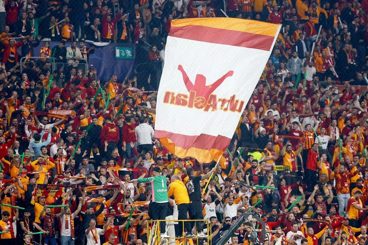 Tłum kibiców tureckich klubów piłkarskich, głównie Galatasaray, świętuje na stadionie, machając flagami i szalikami.