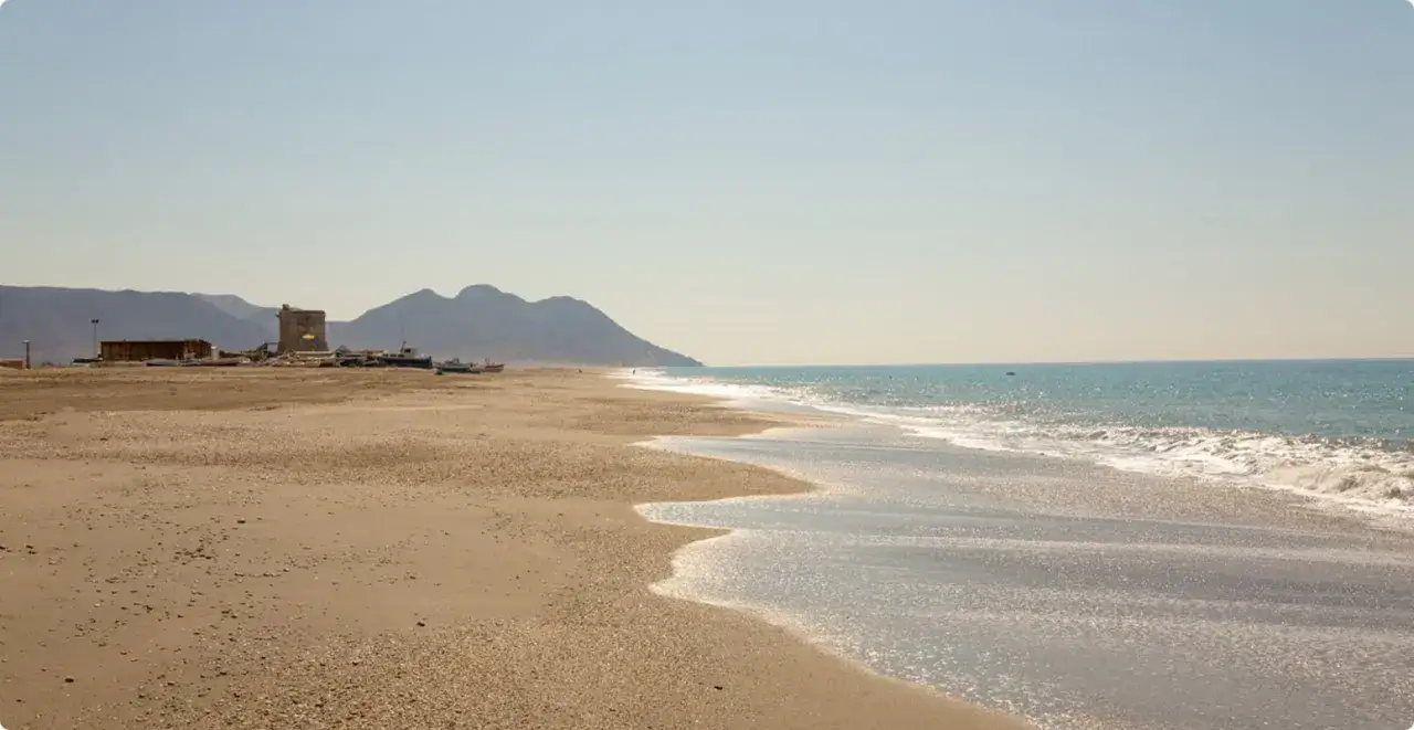 La playa de San Miguel, con sus aguas turquesas y arena dorada, se extiende hacia monta&ntilde;as &aacute;ridas. Barcos y una torre antigua salpican el paisaje.