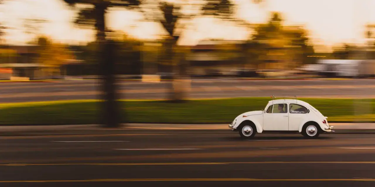 Une Coccinelle blanche roule sur une route, le paysage flou par un effet de photo fil&eacute;. Le soleil couchant illumine la sc&egrave;ne.