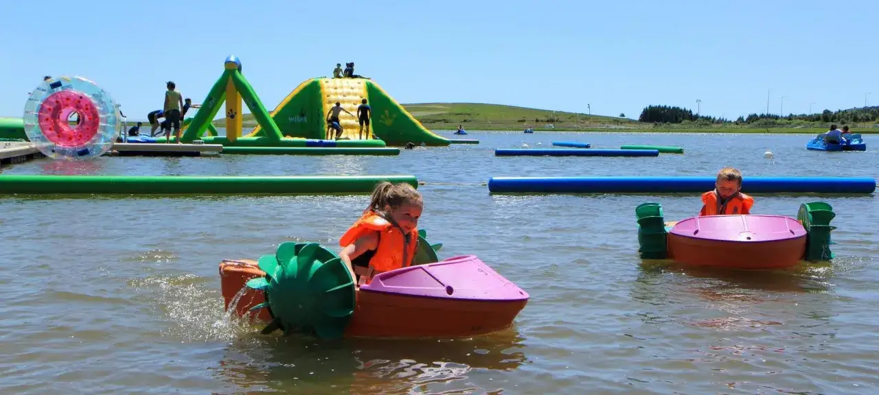 Enfants en bateaux pédalos sur un lac, avec structures gonflables et jeux aquatiques. Idéal pour savoir que faire à Super Besse sans neige !