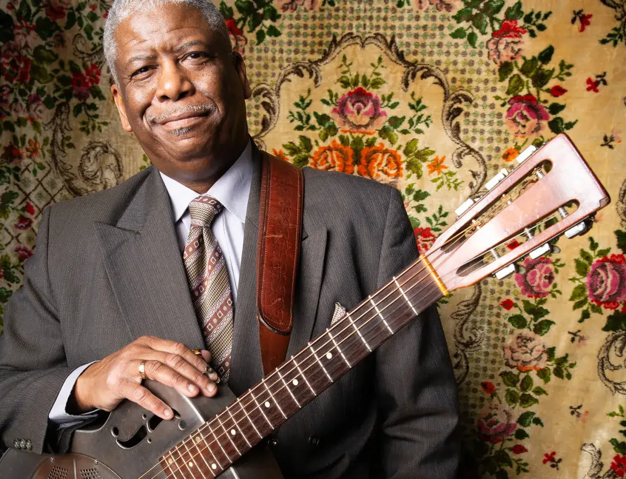 Un musicien souriant, costume gris, cravate à motifs, tient une guitare. Le fond fleuri évoque la naissance du blues.