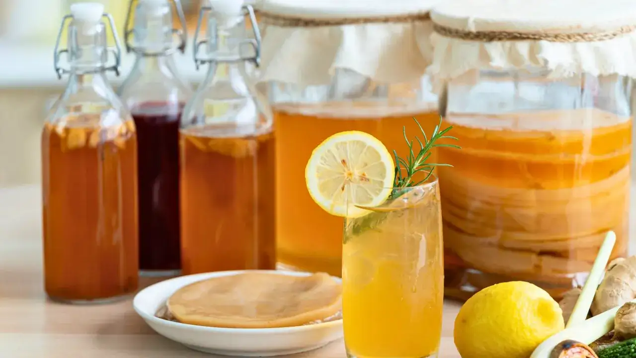 Szklanka orzeźwiającej kombuchy z cytryną i rozmarynem, obok słoje z napojem i SCOBY. Czy kombucha jest zdrowa? Tak, to fermentowany napój pełen probiotyków.