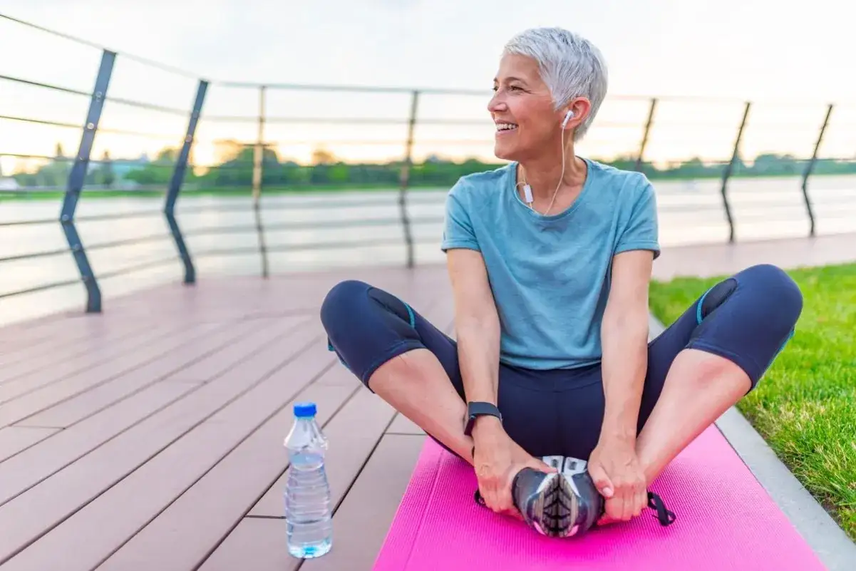 Femme souriante, 50 ans, en tenue de sport, étire ses jambes sur un tapis rose au bord de l'eau. Le sport à 50 ans, c'est la vie !