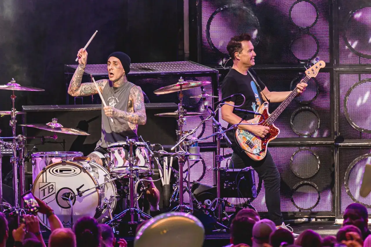Travis Barker trommelt energisch, während Mark Hoppus mit seiner bunten Bassgitarre die Bühne rockt. Mitglieder von blink-182 live in Aktion.
