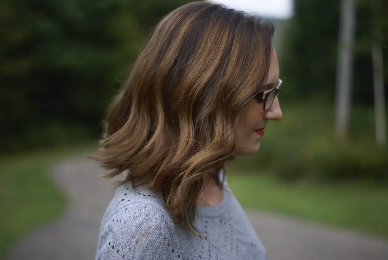 Profil d'une femme aux cheveux châtains avec des mèches brunes sur cheveux blancs, portant des lunettes et un pull gris.