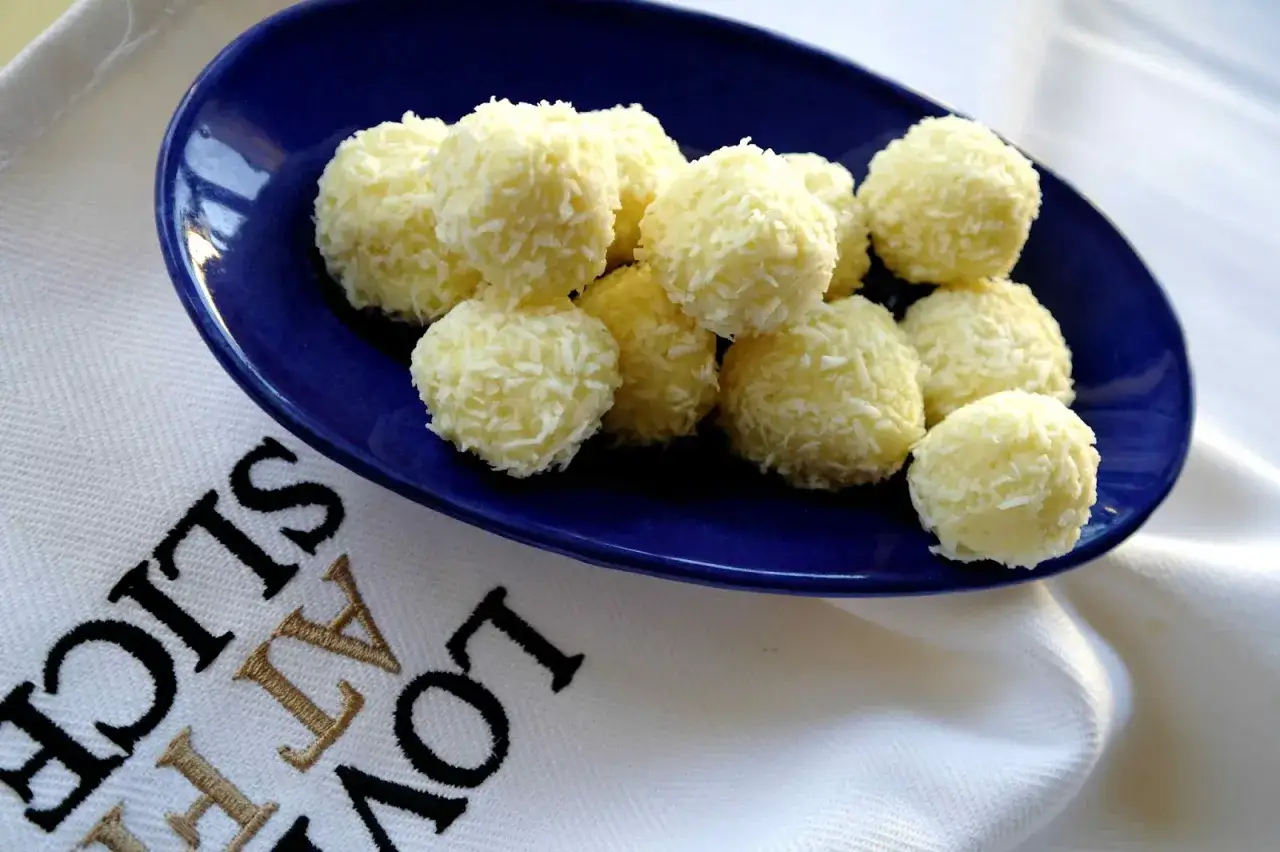 En blå tallrik fylld med fluffiga, vita chokladbollar täckta av kokosflingor. En vit duk med texten