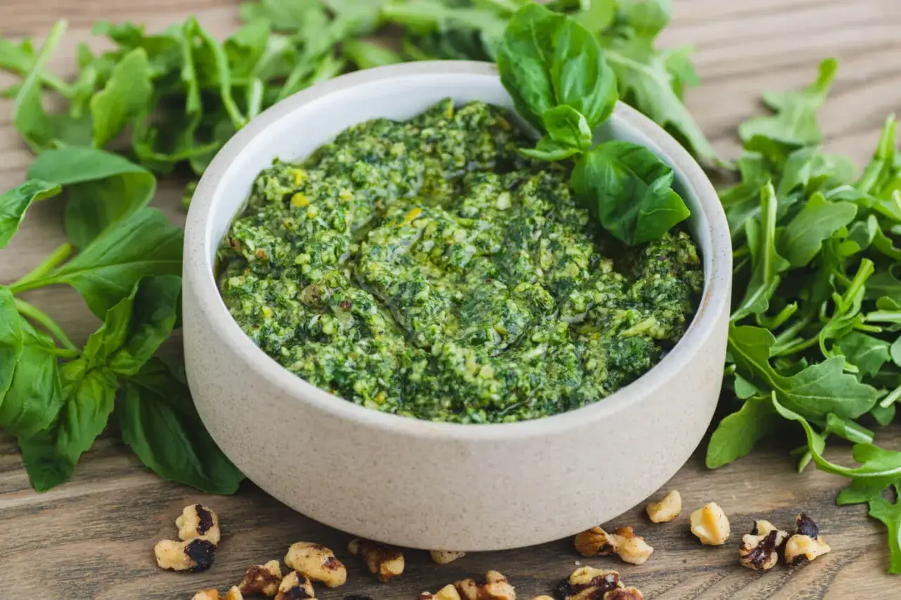 Sch&uuml;ssel mit frischem Rucola Pesto, umgeben von Rucola-Bl&auml;ttern und Waln&uuml;ssen auf einem Holztisch.