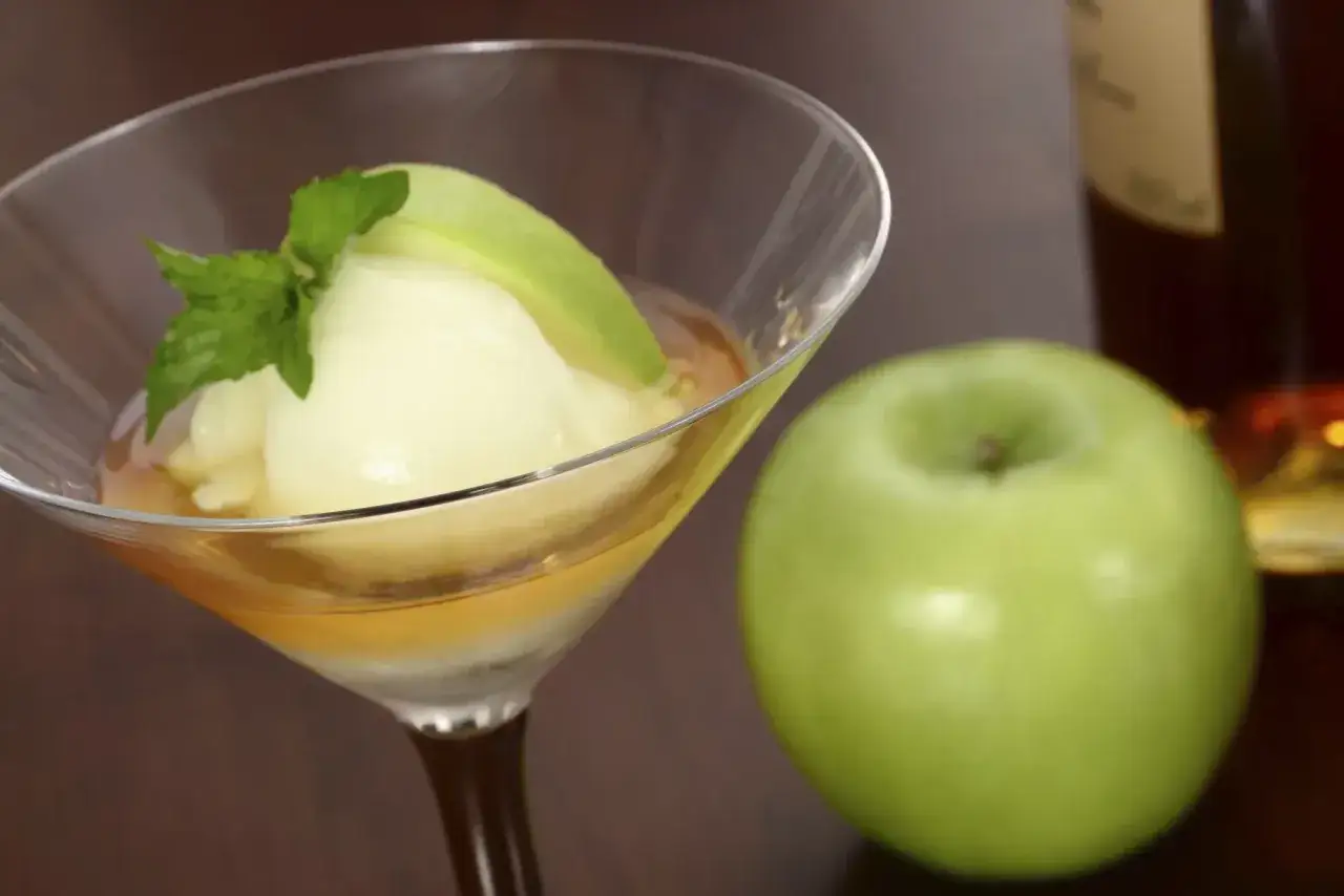 Un trou normand recette : glace à la pomme, calvados et menthe dans un verre à cocktail, avec une pomme verte et une bouteille en arrière-plan.