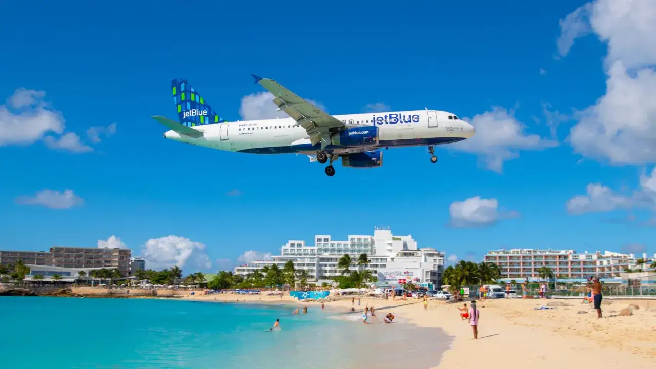 Samolot JetBlue przelatuje nisko nad plażą, tuż przed lądowaniem na lotnisku przy plaży. W tle białe budynki hotelowe i turkusowe morze.
