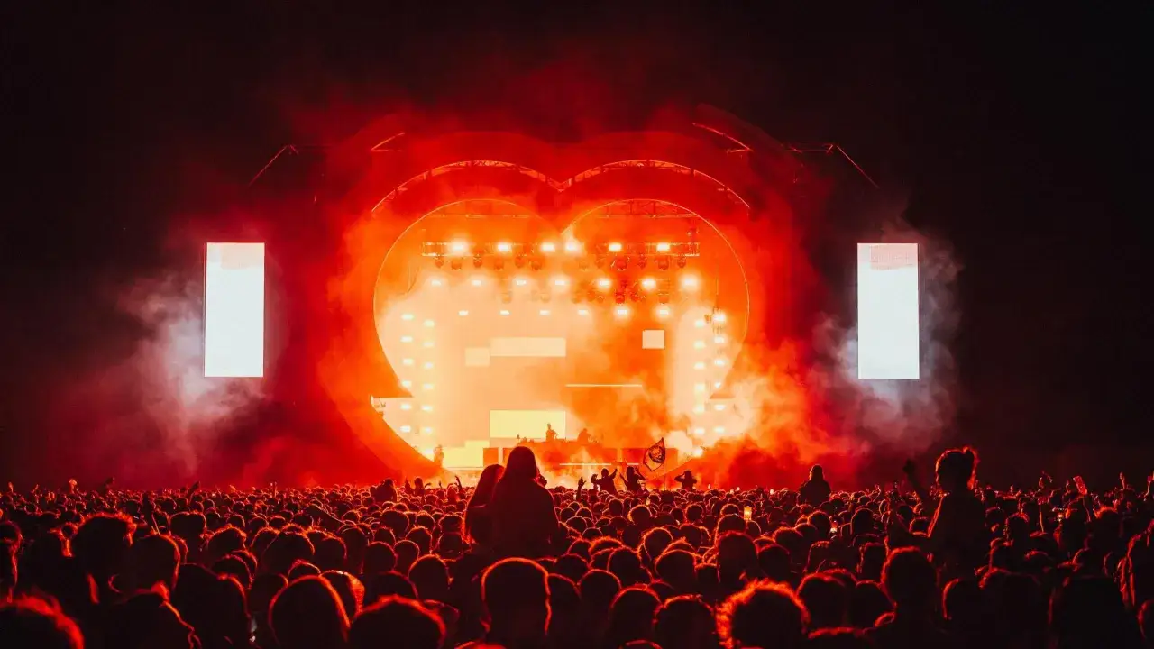 Un concerto con un palco a forma di cuore illuminato da luci rosse e fumo. La folla esulta, immersa nell'atmosfera di una canzone d'amore.
