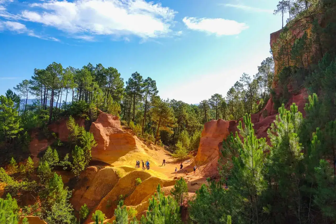 Sentier des ocres, un paysage coloré de falaises rouges et jaunes, parsemé de pins. Des visiteurs explorent ce site naturel unique, rappelant un musée à ciel ouvert.
