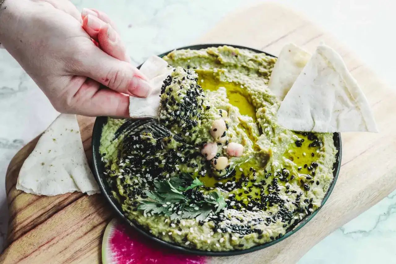 Hummus de aguacate cremoso, adornado con semillas de s&eacute;samo y garbanzos, listo para ser untado con pan de pita.