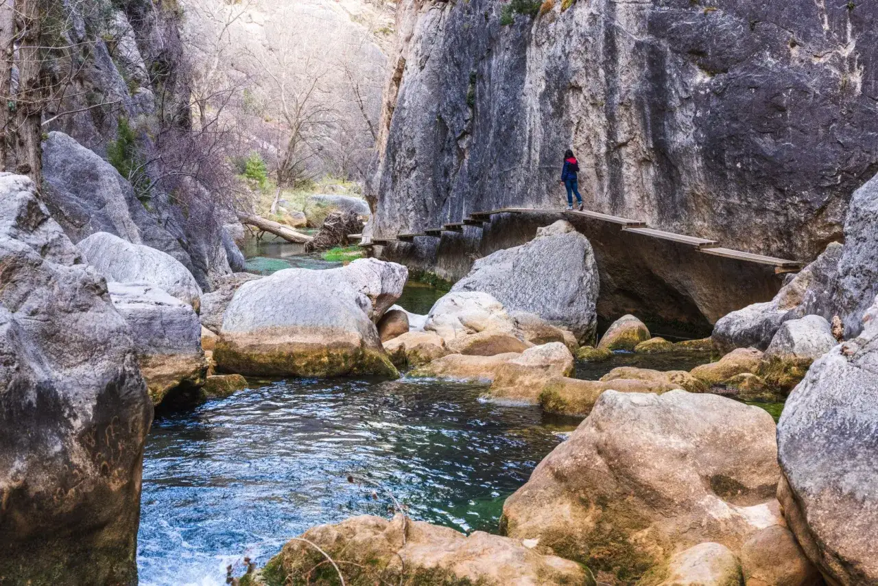 Persona camina por un puente de madera en la ruta del silencio Teruel, rodeada de rocas y un río de aguas cristalinas.
