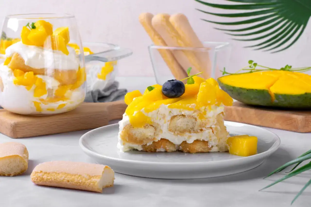 Tranche de tiramisu mangue crémeux, garnie de fruits frais et d'une myrtille. Des biscuits à la cuillère et une moitié de mangue complètent la scène.