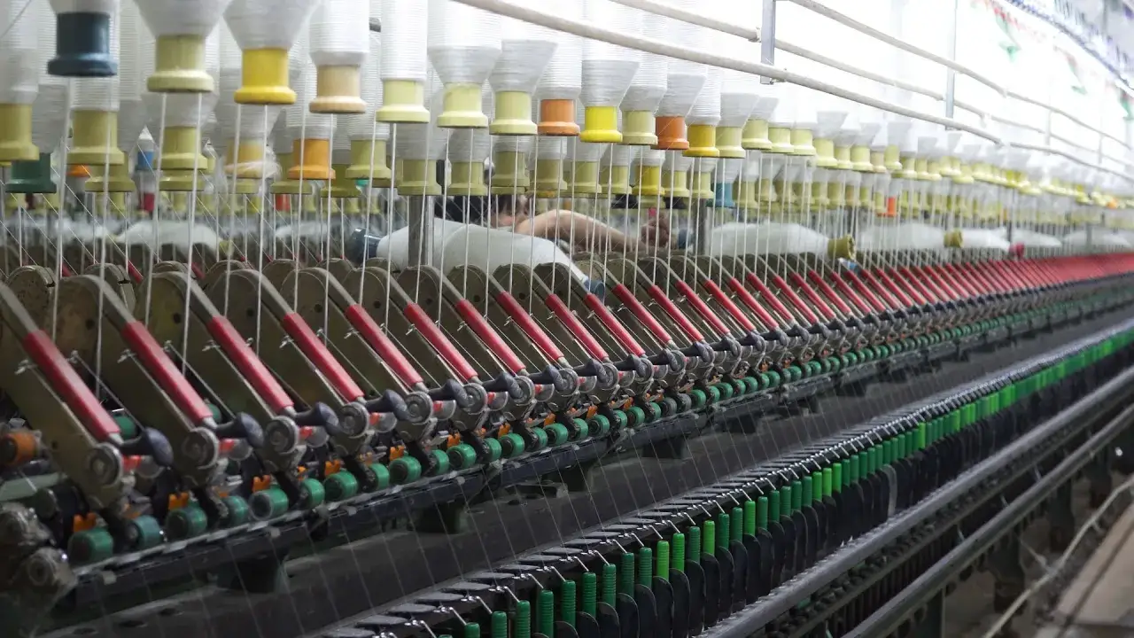 Lignes de machines de filature, avec des bobines de fil blanc et coloré. Les bras mécaniques rouges s'alignent sur des centaines de fils.