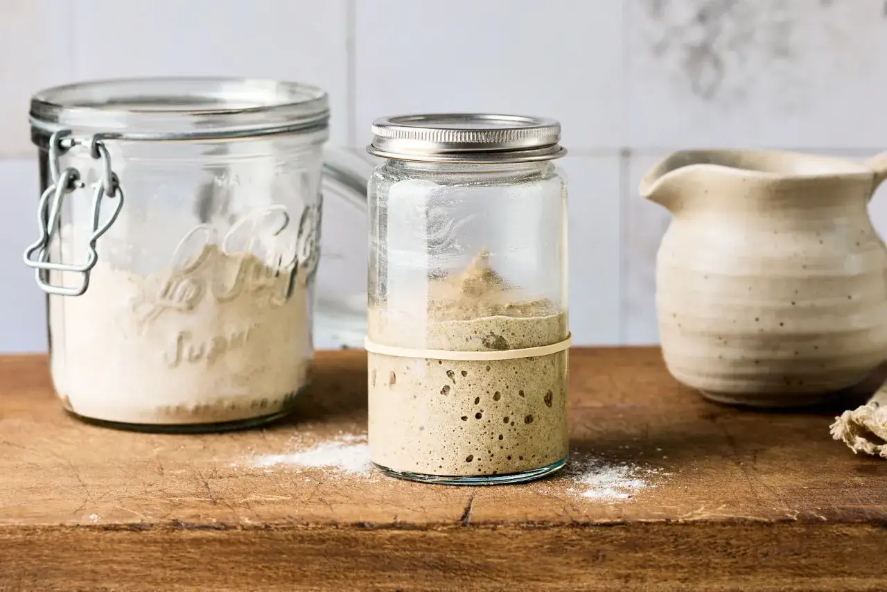 Ein Glas mit Blasen und Aktivität zeigt, wie man Sauerteig ansetzt. Daneben ein weiteres Glas mit Mehl.