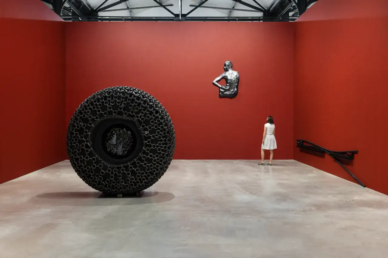 A woman observes a large tire covered in chains and a torso sculpture on a red wall, part of Arthur Jafa art.